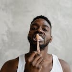 A young black man in a white tank top brushing his teeth, promoting dental hygiene and self-care.