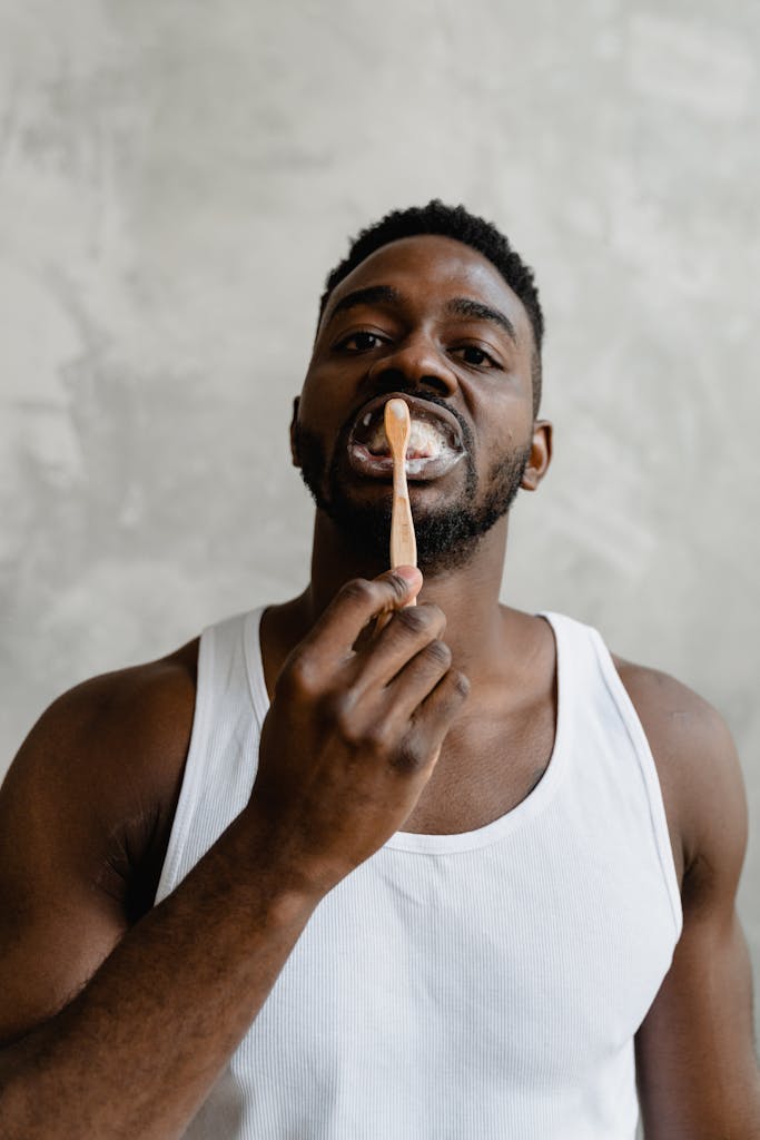 A young black man in a white tank top brushing his teeth, promoting dental hygiene and self-care.