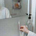 A reflective bathroom scene showing a person's hand reaching for a toothbrush inside a glass cup.