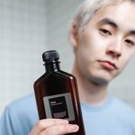 Asian man showcasing skincare tonic bottle for beauty routine indoors.