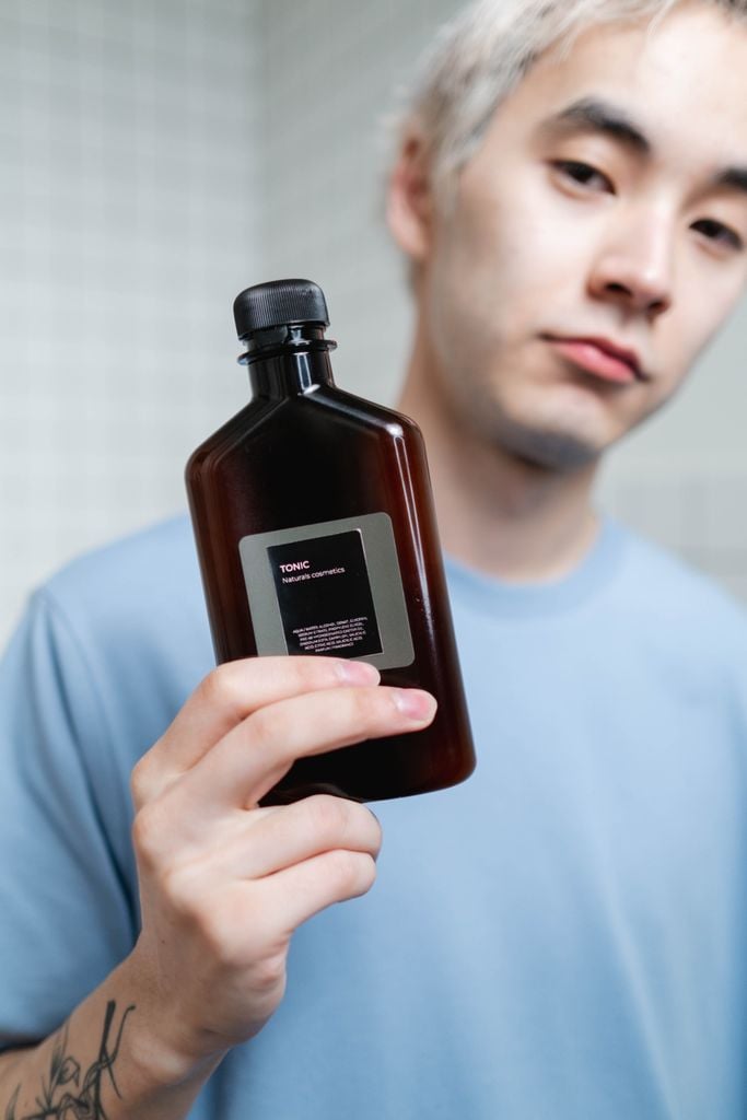Asian man showcasing skincare tonic bottle for beauty routine indoors.